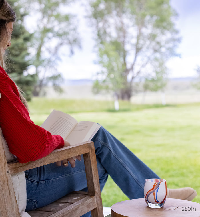 Glass candle holder on table with person reading a book outdoors.