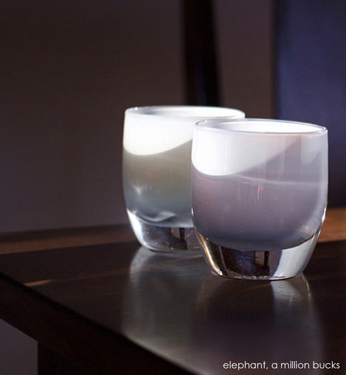 a million bucks light grey hand-blown glass votive candle holder. Paired with elephant sitting in sunlight on dark wood table.