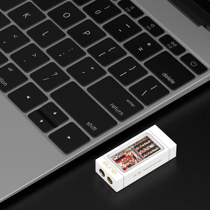 An Aune Audio YUKI Portable DAC and Headphone Amplifier by Aune Audio, featuring a white casing with gold ports and visible circuit components, lies next to a laptop keyboard on a black surface. This compact device delivers high-quality audio while serving as both a powerful headphone amplifier and a portable USB DAC.