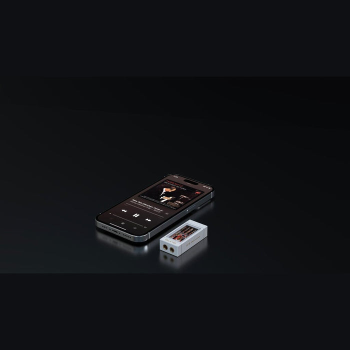 A smartphone playing music is placed next to the Aune Audio YUKI Portable DAC and Headphone Amplifier by Aune Audio—a small, rectangular audio device with a screen, headphone jack, and portable USB DAC functionality—both resting on a dark, reflective surface.