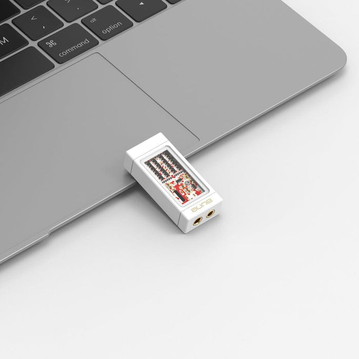 An Aune Audio YUKI Portable DAC and Headphone Amplifier by Aune Audio, featuring a compact white design with visible circuitry, is plugged into a silver laptop on a white surface. The laptop's keyboard and device ports are partially visible.