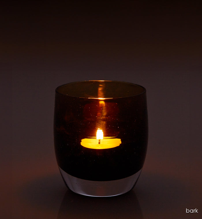 bark, transparent rich dark brown, hand-blown glass votive candle holder with a lit candle on a dark background