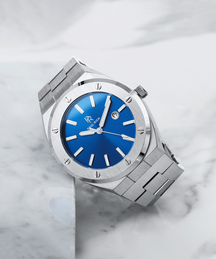 Paul Rich brushed silver watch with vivid blue sunray dial and date window, placed diagonally on white marble surface.