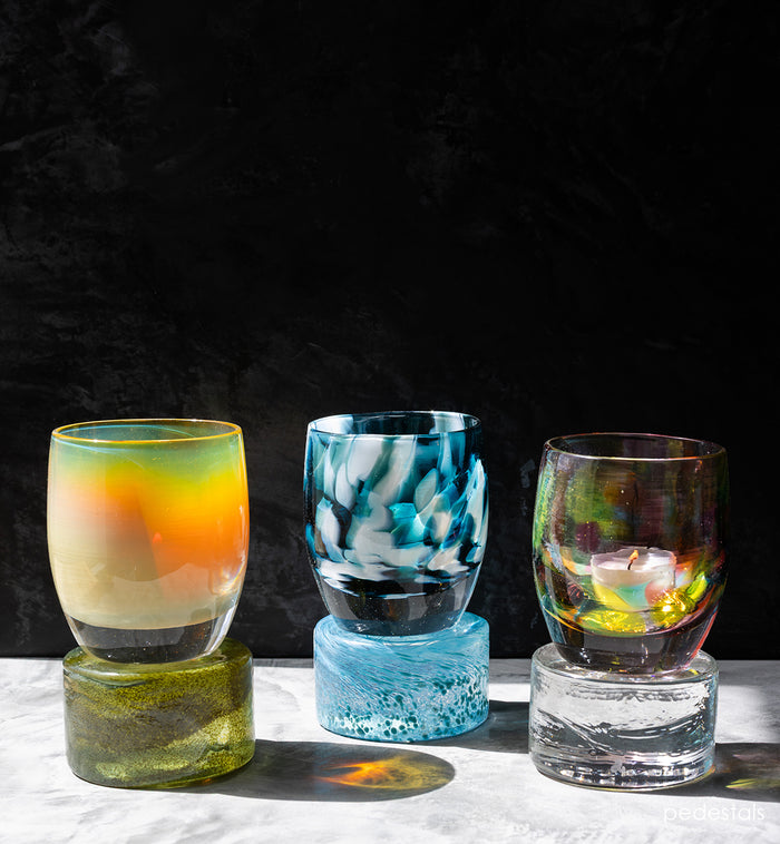Three colorful glass pedestals holding lit glassybaby on a dark background