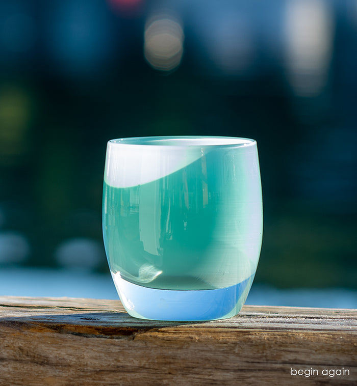 begin again light teal, hand-blown glass votive candle holder sitting in sunlight on wood perch with a body of water and boat blurred in background.