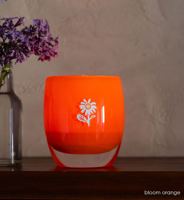 bloom orange glassybaby with white flower etched on sitting on wood shelf.