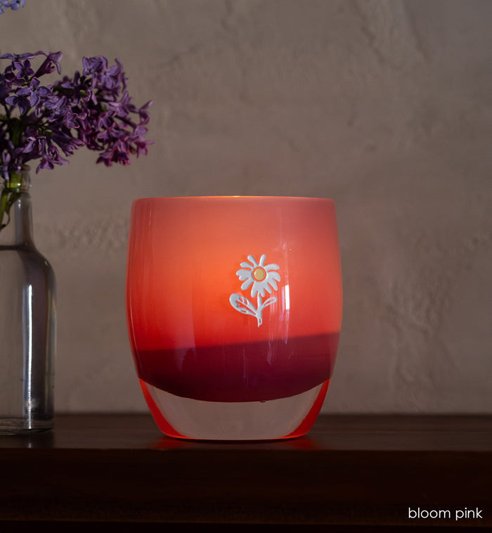bloom pink glassybaby with white flower etched on it sitting on wood shelf.