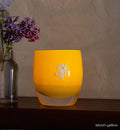 bloom yellow glassybaby with white flower etched on it sitting on wood shelf.