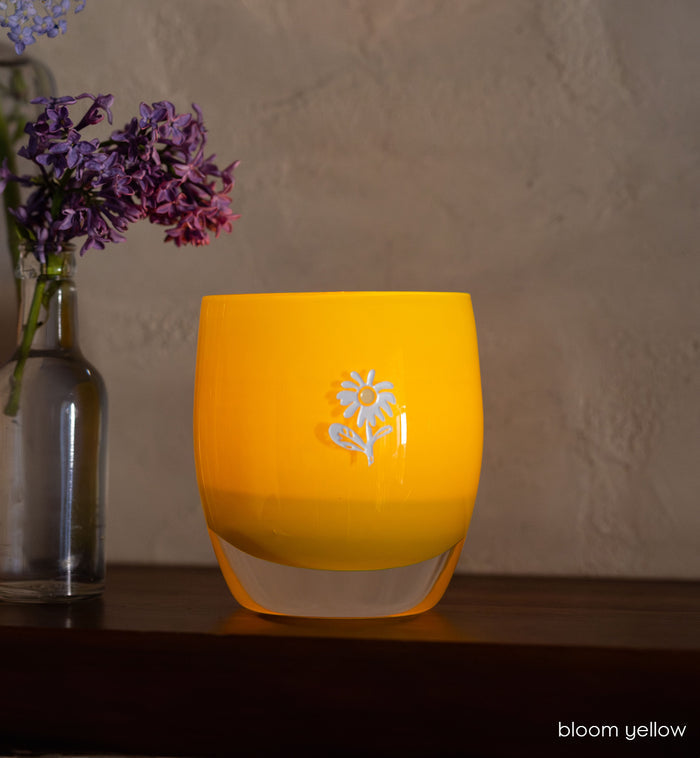bloom yellow glassybaby with white flower etched on it sitting on wood shelf.