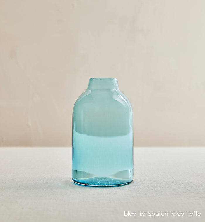 blue transparent bloomette, light transparent blue hand-blown glass flower bud vase in front of cream background.