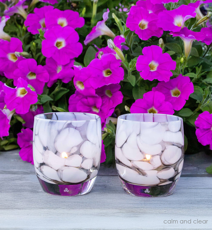 two calm and clear gray and white petal, hand-blown glass votive candle holders on gray table with purple flowers.