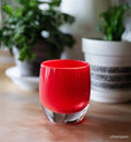 Red hand-blown glass candle holder on a wooden surface with blurred plants in the background