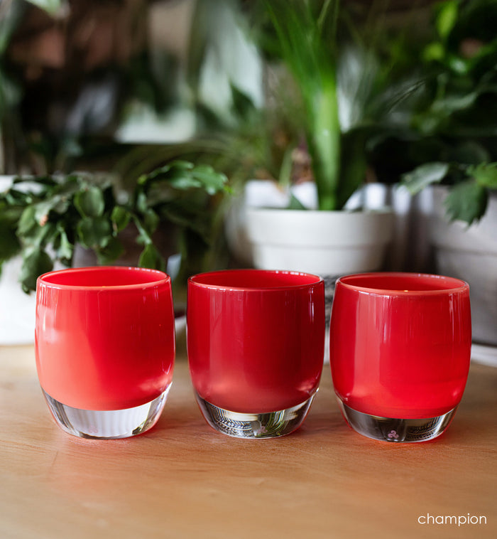 Three red hand-blown glass candle holder on a wooden surface with blurred plants in the background