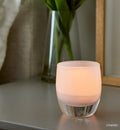 cherish soft white hand-blown glass votive candle holder on a gray nightstand with a vase and mirror.