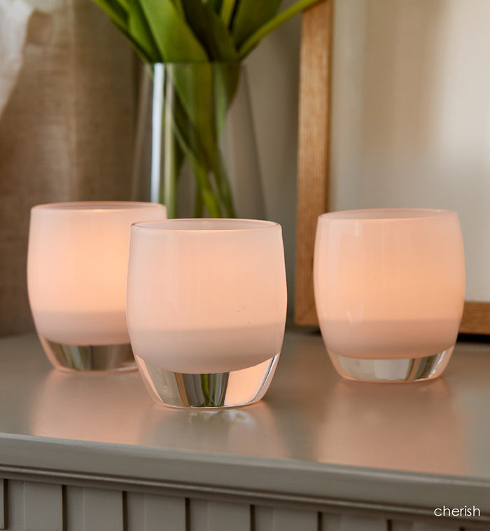 three cherish soft white hand-blown glass votive candle holders on a gray nightstand with a vase and mirror.