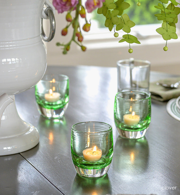 three clover, transparent light green hand-blown glass votive candle holders on a dining room table with a large vase of flowers and diningware.