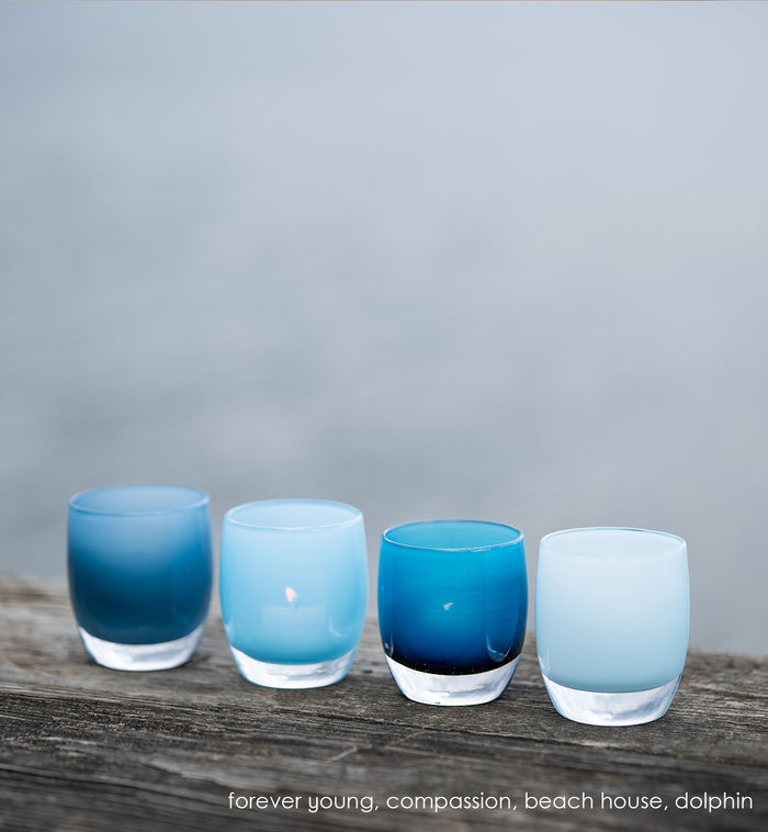 compassion powder blue, hand-blown glass votive candle holder in a line up with forever young, beach house and dolphin outside on wood surface.