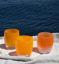 cozy, stripey apricot orange hand-blown glass votive candle holder with sassy and cozy on a beach towel in front of a body of water.