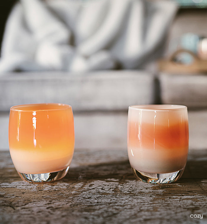 cozy, two apricot orange stripey hand-blown glass votive candle holders on a wood coffee table with a gray cozy couch in background.