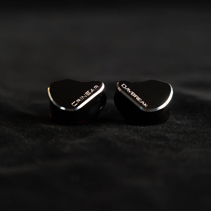 Two black Crinear Daybreak In-Ear Headphones, labeled “CRIN EAR” and “DAYBREAK,” rest on a dark surface with reflective metal trim—crafted for audiophile sound and available at headphones.com. Brand: Crinear.