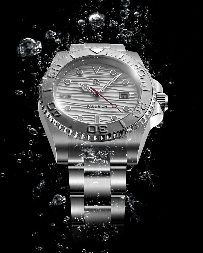 Stainless steel watch with a silver wave-textured dial shown against splashing water on a black background, highlighting the bezel, bracelet, and red seconds hand.