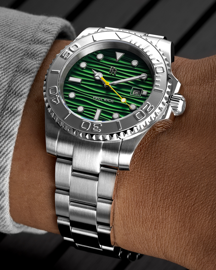 Stainless steel watch with a green wave-textured dial and yellow seconds hand shown on a wrist, highlighting the bezel, markers, and brushed bracelet.