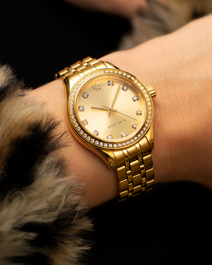 Gold-tone Paul Rich watch with gold sunray dial, diamond hour markers, and gem-set bezel, worn on a wrist with a faux fur coat sleeve.