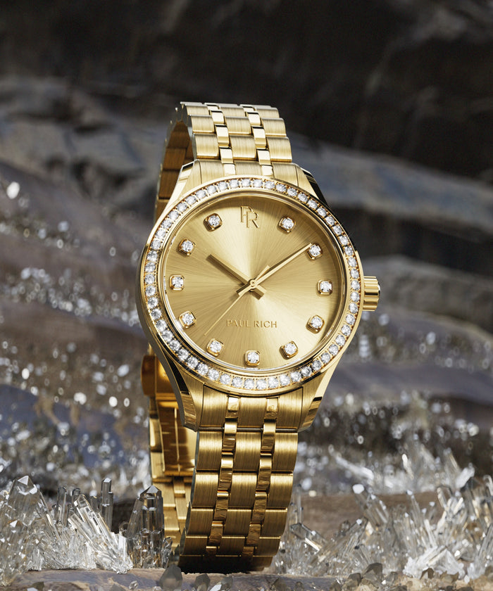 Gold-tone Paul Rich watch with sunray gold dial, diamond hour markers, and gem-set bezel, displayed upright on a bed of clear crystals.