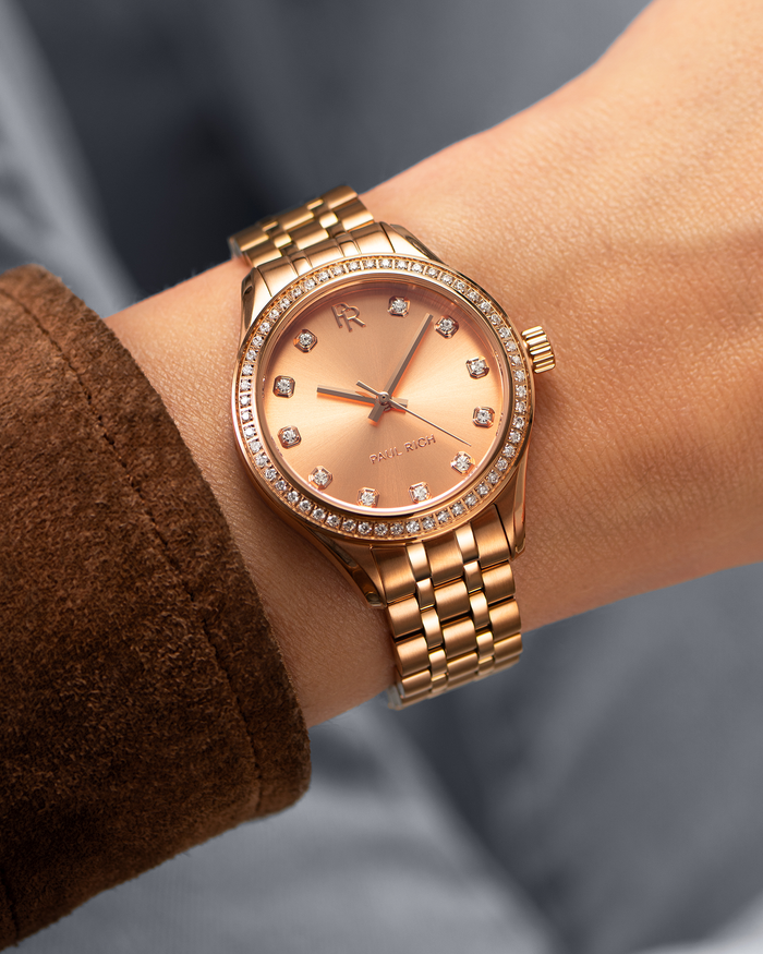 Rose gold Paul Rich watch with a matching sunray dial, diamond hour markers, and gem-set bezel, worn on wrist with a brown suede sleeve.