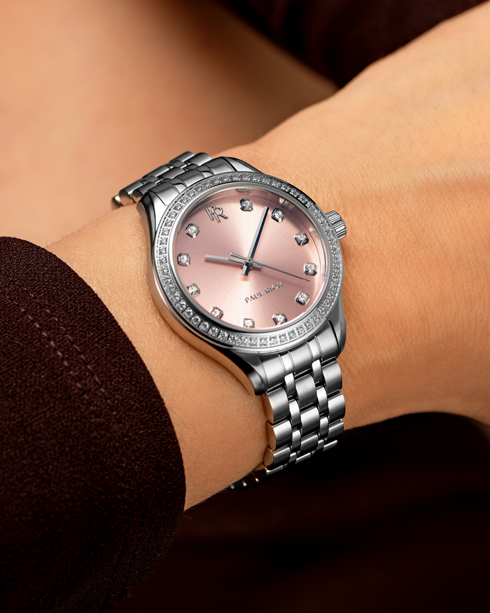 Silver-tone Paul Rich watch with pink sunray dial, diamond hour markers, and gem-set bezel, worn on wrist with a dark burgundy sleeve.