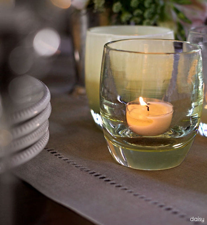 daisy transparent crystal yellow hand-blown glass votive candle holder placed on a table runner on a dining room table next to ceramic plates.
