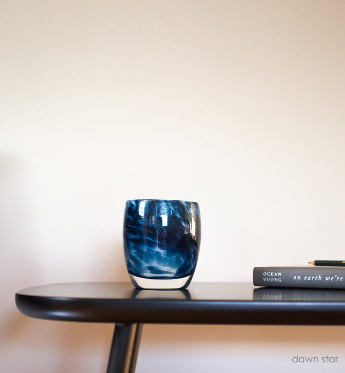 dawn star blue glitter, hand-blown glass votive candle holder on a black indoor bench next to a book and pencil.