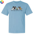 A light blue short-sleeve crewneck t-shirt featuring a graphic of four different dog breeds sitting next to a rainbow-colored book, with the text 'don't judge a bark by its size' printed below. A small multicolor color wheel icon is visible in the top-left corner.