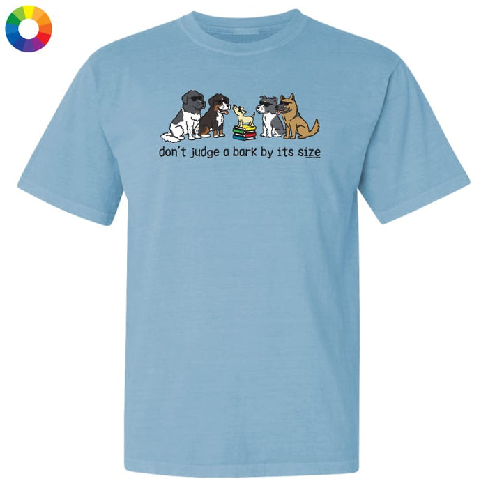 A light blue short-sleeve crewneck t-shirt featuring a graphic of four different dog breeds sitting next to a rainbow-colored book, with the text 'don't judge a bark by its size' printed below. A small multicolor color wheel icon is visible in the top-left corner.