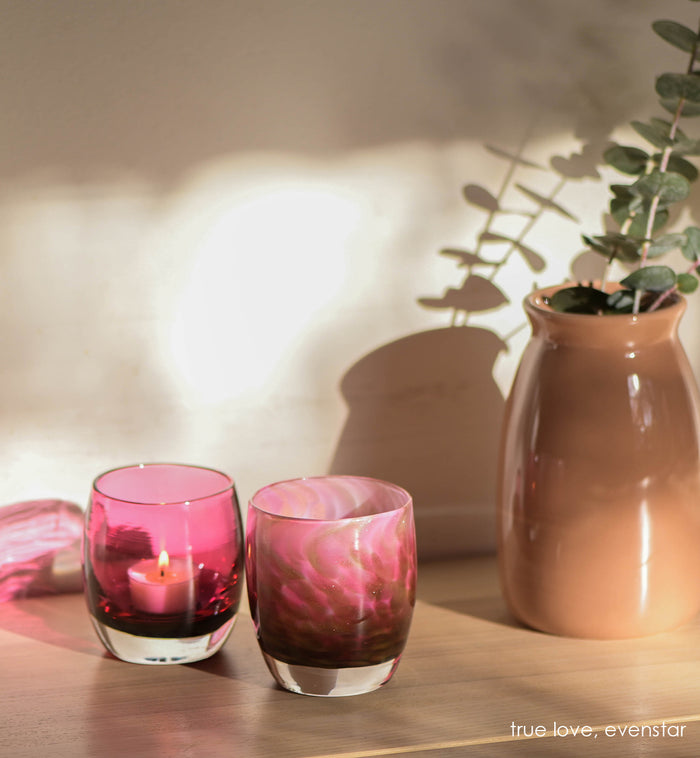 evenstar pink and gold glitter hand-blown glass votive candle holder with true love on a wood table to a potted plant.