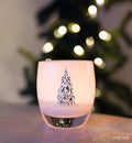 white with silver etched tree, hand-blown glass votive candle holder a lit Christmas tree in the background