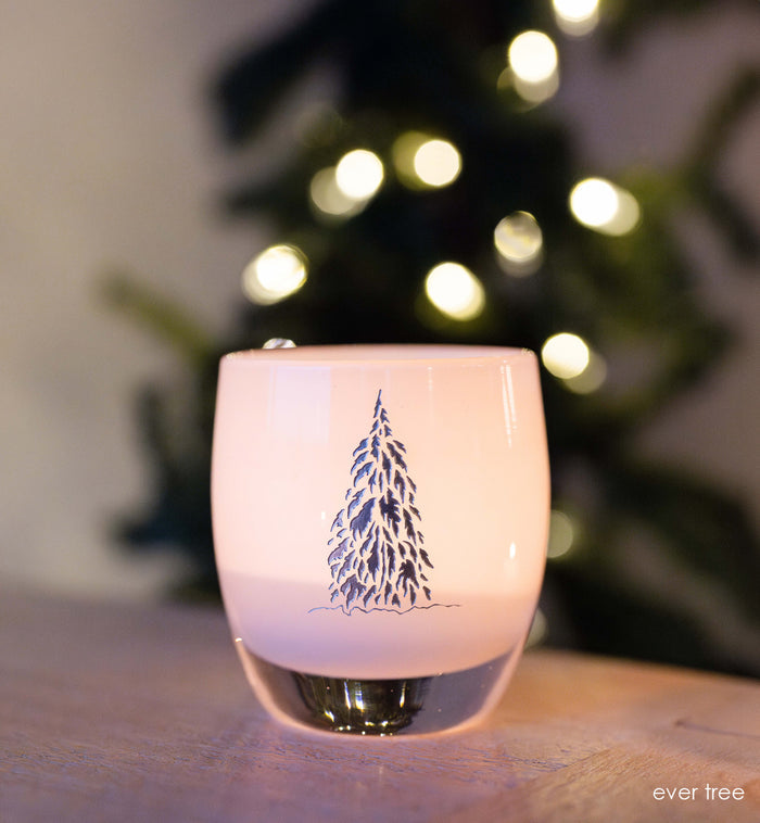 white with silver etched tree, hand-blown glass votive candle holder a lit Christmas tree in the background