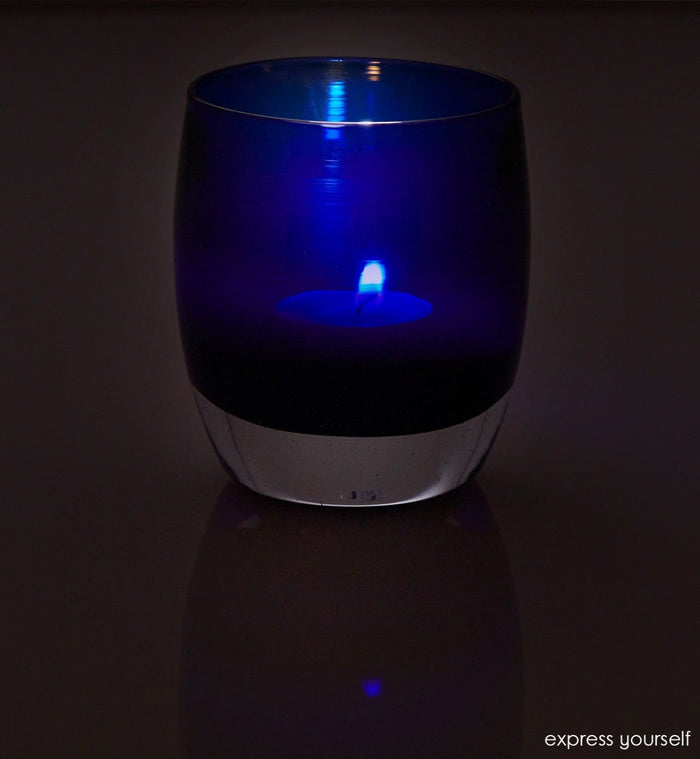 express yourself, transparent admiral blue, hand-blown glass votive candle holder lit in a dark room against a reflective surface.