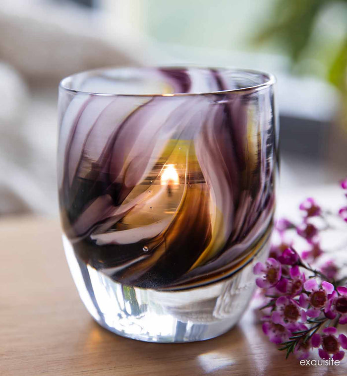 exquisite, purple gray yellow swirl, hand-blown glass votive candle holder on a light wood surface with purple flowers.