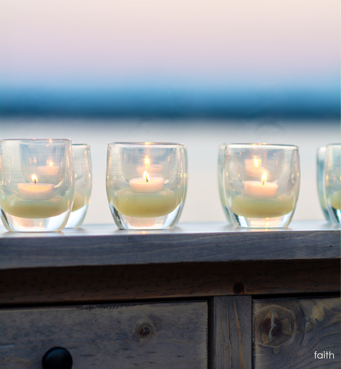 several faith translucent cream hand-blown glass votive candle holders on a wood dresser overlooking ocean water and the sunset.