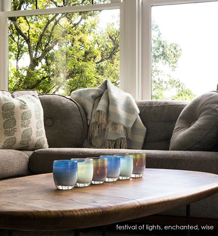 festival of lights, light and dark blue with metallic luster hand-blown glass votive candle holder with wise and enchanted on a wood coffee table next to a gray couch.
