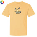 A light peach-yellow short-sleeve t-shirt with a cartoon graphic of a dog sitting on a beach towel with seagulls flying around and the text 'flock off' underneath. A small multicolor color wheel icon is in the top-left corner.