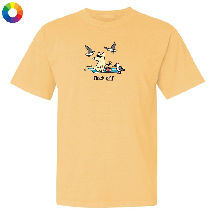 A light peach-yellow short-sleeve t-shirt with a cartoon graphic of a dog sitting on a beach towel with seagulls flying around and the text 'flock off' underneath. A small multicolor color wheel icon is in the top-left corner.