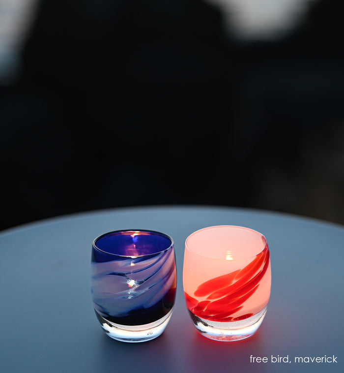freebird blue with white wing next to maverick white with red wing, hand-blown glass votive candle holders on a dimly lit table.