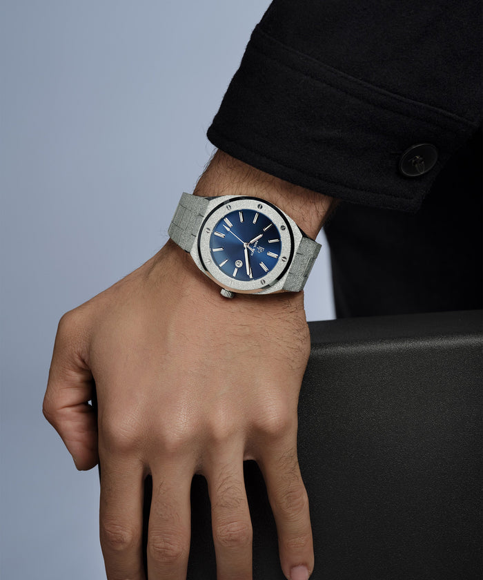 Hand wearing a watch with a blue face and gray strap on a neutral background