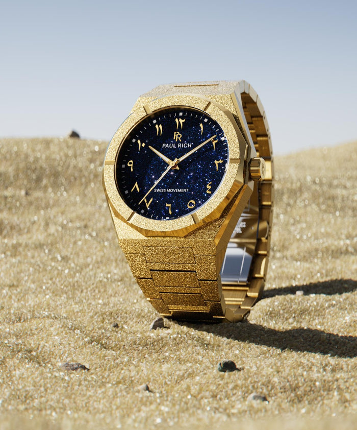 Frosted gold watch with a blue aventurine dial and Arabic numerals, standing upright on sunlit sand with a clear blue sky in the background.