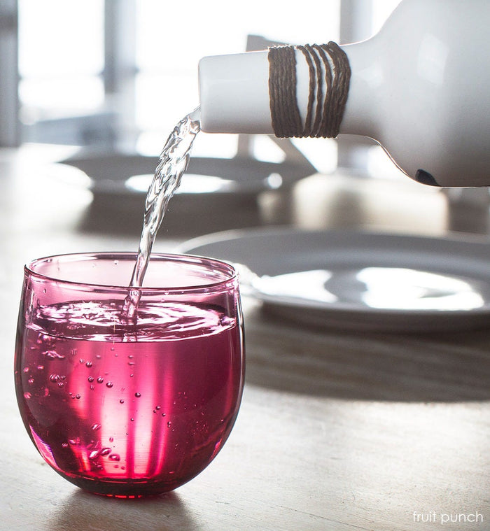 fruit punch drinker, transparent raspberry, hand-blown drinking glass, having bubbly water poured in on a light wood dining table.