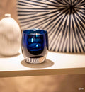 give, hand-blown sapphire blue with silver metallic interior, hand-blown glass votive candle holder lit with a tealight on a creams shelf with a vase and piece of art.