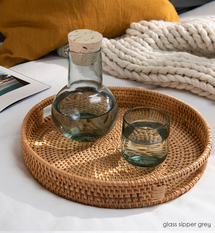 glass sipper grey translucent dark grey hand-blown decanter sitting on a wicker tray filled with water on a cozy couch surrounded by a blanket, pillow, and book.