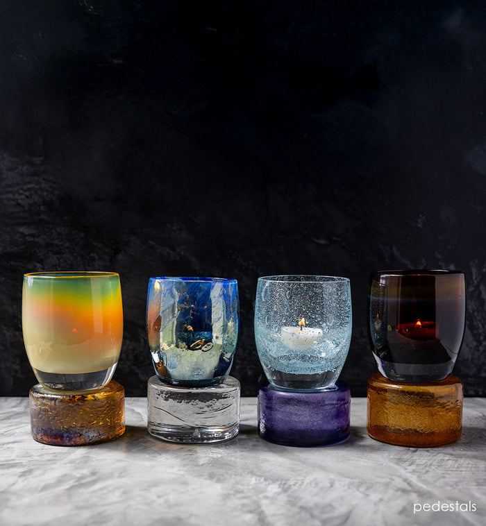 Five colorful glass candle holders on pedestals against a dark background